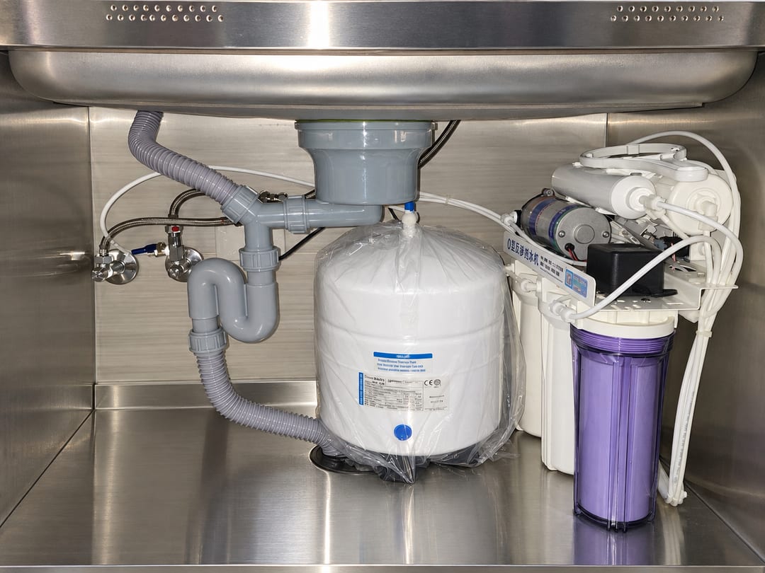 不鏽鋼廚櫃裡的有壓力桶 RO 機：左邊白色壓力桶，右邊三道濾心與紫色濾瓶